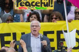A white-haired white man with glasses and wearing a blazer is speaking and holding a microphone. Around him is a crowd of people holding cameras, protest signs and a banner.