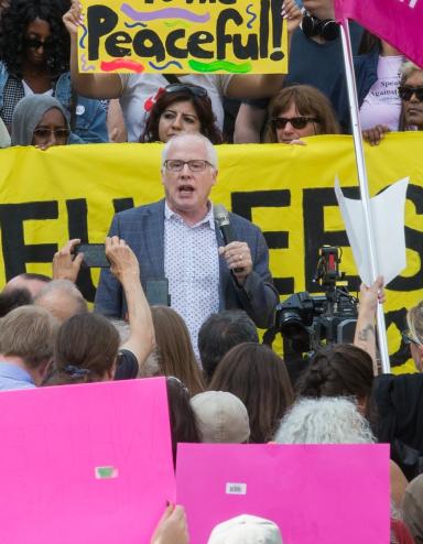 A white-haired white man with glasses and wearing a blazer is speaking and holding a microphone. Around him is a crowd of people holding cameras, protest signs and a banner. Partially obscured.