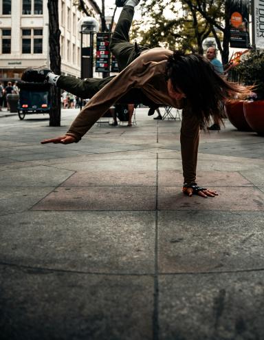 A woman in a grey jacket and darker grey pants is breakdancing on a sidewalk during daytime. Trees and buildings are in the background. Partially obscured.