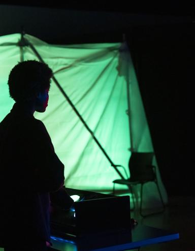 The silhouette of a person standing in the dark in front of red and green clothed box structures lit from the inside and containing shadowed figures. Partially obscured.