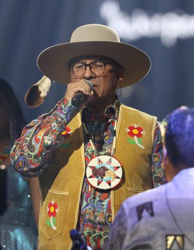 Close-up on a group of people wearing beaded jackets and vests performing on stage. In the centreer, a man wearing glasses, a hat, and a large, beaded medallion sings into a microphone. Partially obscured.