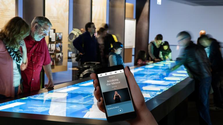 A hand holds up a mobile phone. On the phone are the words “Floor 4” and “Breaking the Silence,” and a person speaking using sign language. In the background, Museum visitors are looking at a large rectangular table with a touchscreen. Partially obscured.