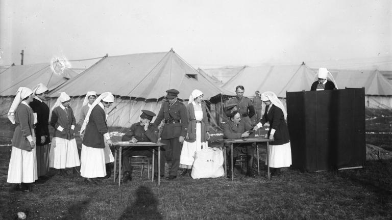 A black-and-white photograph of nurses and men in soldiers’ uniforms in front of a set of tents. To the right of the image, a nurse stands behind a voting screen. Only her head and shoulders can be seen.
