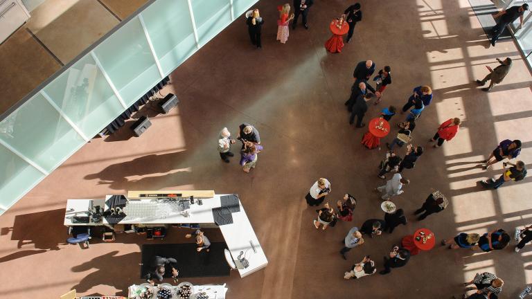 A bird’s-eye view of a crowd of people gathered in a large open area in the Museum. There are cocktail tables with red tablecloths and a bar on the left side of the image. Partially obscured.