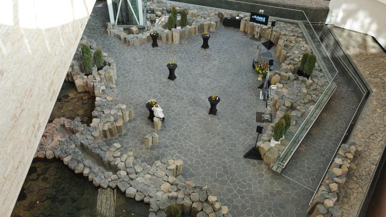 A view from above of an area surrounded by light brown or grey stones. Five cocktail tables with black tablecloths are set up.