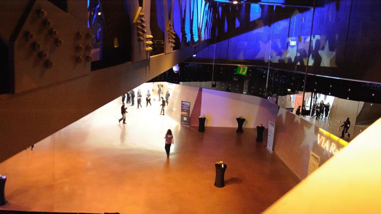 A view looking down on a large open space with visitors socializing in the hall below. Blue light and stars are projected onto the far wall. Partially obscured.