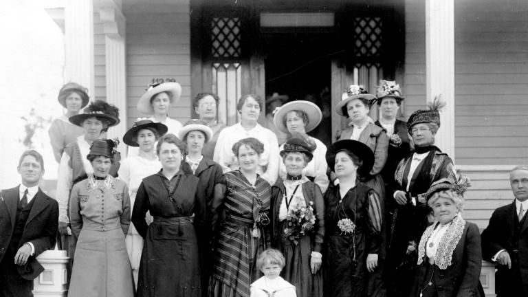 A black-and-white photograph of 17 women, two men and a child posing on the front steps of a house. All are dressed in early twentieth-century clothing.