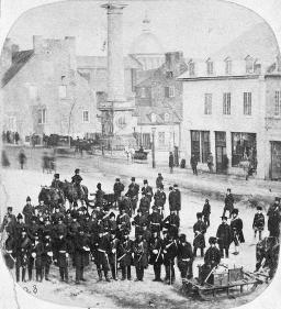 A black-and-white photo of a large number of soldiers in 19th century uniforms standing along a street with buildings made of stone and wood.