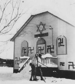 Two people on skis in front of a synagogue with several swastikas painted on it.