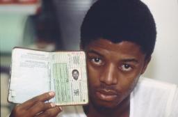 A young Black man faces the camera, holding up a booklet resembling a passport.
