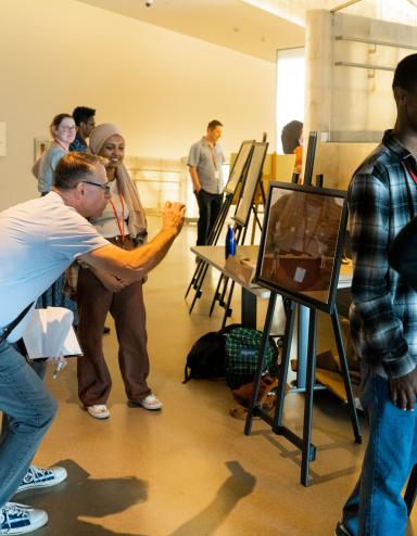 In a museum gallery, students exhibit projects on easels. Two students speak with adults about their work. Partially obscured.