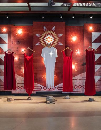 Six red dresses and a white dress in the middle are suspended on wooden clothes hangers in front of a backdrop. A white star glows at the location of the heart on the central white dress. The backdrop features patterns on a birchbark texture. Some of the patterns are illuminated from behind to create glowing star-like features on the panels. Partially obscured.