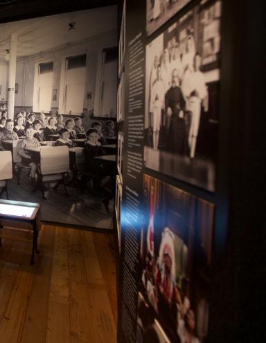 A Museum exhibit showing a black-and-white photo of children sitting in rows at school desks. Two desks, similar to those in the photo sit in the centre of the exhibit. A headline on a text panel reads “Childhood Denied.” Partially obscured.
