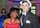 Two young people pose for a head and shoulders photograph. On the left is a young Black woman wearing a red strapless dress. On the right is a young white man in a black tuxedo and a white fedora.