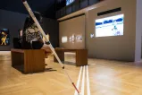In the foreground, a visually impaired person’s white stick touches three tactile strips laid out on the floor of a Museum gallery. The strips lead to benches, where two people are watching a film on a large screen.