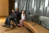 An older man sits in a wheelchair beside a young girl sitting on a bench. The man is pointing up, and both he and the young girl are looking up and to the viewer’s left.