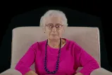 An older lady wearing glasses and a pink shirt sits on a chair with her hands folded in her lap. She is looking straight at the camera with a neutral expression.