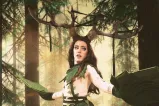 A drag artist is dressed in a cosplay costume with long, brown hair; large antlers; and green leaf scarf, skirt and gloves.