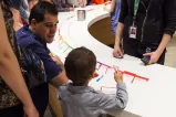Museum visitors of all ages are writing on cards and talking to one another around a curved table. 