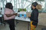 Two women speaking in between a poster that reads "Basics for the blind, Freetown, Sierra Leone"