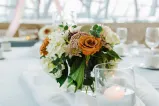Green, white and orange floral centre piece with a dinner table set in the background.