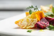 Bright gold-coloured potato wedges are artfully arranged on a square white plate, along with leaf greens, wedges of red beets, and garnish.