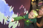 A woman and a young boy look up at a wall. The boy is reaching out and touching the wall. Blue, red and green coloured light is being projected onto the wall and the boy’s hand.