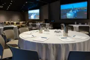 A large classroom is set up for a conference or, meeting with two large screens on the wall, round tables covered with white tablecloths, notepads and pens, and grey plastic chairs.