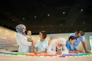 A family of six stands in front of curved table with coloured cards on it. Three family members are writing on cards with markers while the woman holds a smiling toddler.