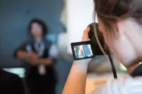 A person looks through the screen of a camera in the foreground while a person poses in the background. 