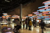 A large number of people in a gallery with a wall of colourful panels and photos showing a timeline of events.