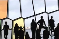 Eighteen people in a row, some seated, some standing, in front of a wall of odd-shaped windows. 
