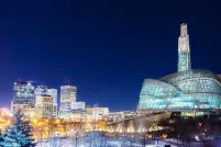 A glass and steel building on a snowy evening, positioned behind an illuminated sign that says Winnipeg.