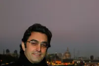 Close-up of a smiling man at dusk. Behind him is a body of water and on the other side there are boats, lights and buildings. 