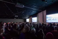 A seated audience viewing three large screens in a dimly lit room.