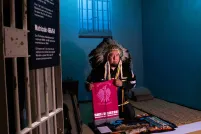An Indigenous man wearing ceremonial regalia kneels inside a replica of a prison cell. He is presenting a textile banner with a graphic image of a bird and the words “bring our children home.” 