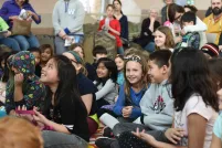 Smiling children sitting cross-legged on the floor. One girl in the centre is looking into the camera.