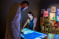 A man and woman look down at an illuminated table. In the background, there is a grouping of colourful posters on the wall.