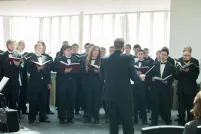A choir of men wearing black outfits hold books and sing in front of a conductor.