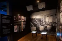 A Museum exhibit showing a black-and-white photo of children sitting in rows at school desks. Two desks, similar to those in the photo sit in the centre of the exhibit. A headline on a text panel reads “Childhood Denied.”