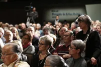 A room full of audience members sitting. One woman is standing and smiling holding a microphone.