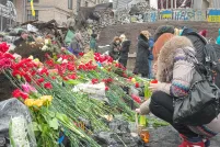 Two people laying red flowers at a memorial. In the background a dozen other people are standing in front of debris, such as rocks, metal sheeting and cement barriers painted with blue and yellow Ukrainian flags and lettering.