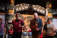 Two men and a woman dance side by side. One man is wearing a red vest and a headdress. The other man, in a suit jacket, carries a bentwood box carved in west coast Indigenous style. In the background are two larger totem poles and people dressed in regalia.