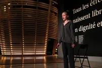 A young woman talks in front of a black wall that says, in large white letters, “Tous les êtres humains naissent libres et égaux en dignité et en droits.” (All human beings are born free and equal in dignity and rights.) There is a circular theatre in the background that resembles a large woven basket.