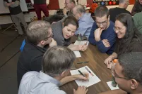 A group of excited people gathered around a table where one man is writing on a piece of paper.