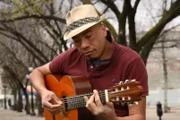 A man wearing a fedora plays a guitar while sitting outside on a bench. There are trees in the background.