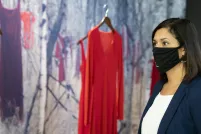 A woman wearing a black face mask stands in front of a museum exhibit consisting of red dresses hanging in front of a woodland background.