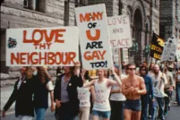 A group of people march down a city street. At the front of the group, on the left side of the image, two people hold a sign over their heads that reads “Love thy neighbour.” Slightly behind them is a sign that reads “Many of u are gay too!”