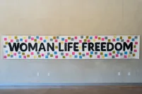 A white banner hangs on a wall, bearing the words "Woman, Life, Freedom" in black. Small coloured squares with text are attached to the banner around the text.