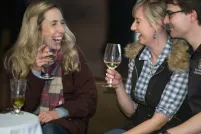 Three laughing people sit together at a table in a Museum space, drinking wine.
